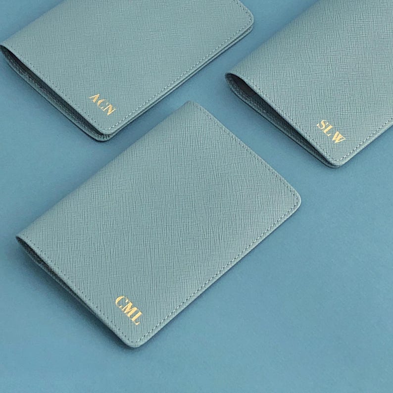May include: Three light blue passport holders with a textured crosshatch finish. Each rectangular cover features gold foil embossed initials in the bottom corner, specifically the letters ACN, SLW, and CML, arranged in a clean, simple layout.
