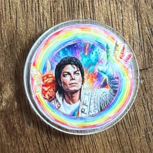 May include: A circular acrylic pin featuring a colorful illustration of Michael Jackson with a small orange creature on his shoulder. The design is surrounded by a vibrant rainbow arc against a cosmic, nebula-like background with stars and bright colors.