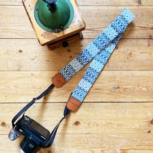 May include: A blue and white patterned camera strap with brown leather accents. The strap is attached to a black camera.
