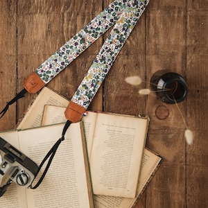 Peut inclure: Un sangle de caméra à motif floral avec des extrémités en cuir marron. La sangle est posée sur une surface en bois avec une pile de livres et une bouteille en verre.