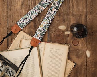 May include: A floral patterned camera strap with brown leather ends. The strap is lying on a wooden surface with a stack of books and a glass bottle.
