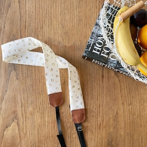 Può includere: Un cinturino per fotocamera bianco con un motivo floreale e dettagli in pelle marrone. Il cinturino è attaccato a una fotocamera bianca con un obiettivo nero.