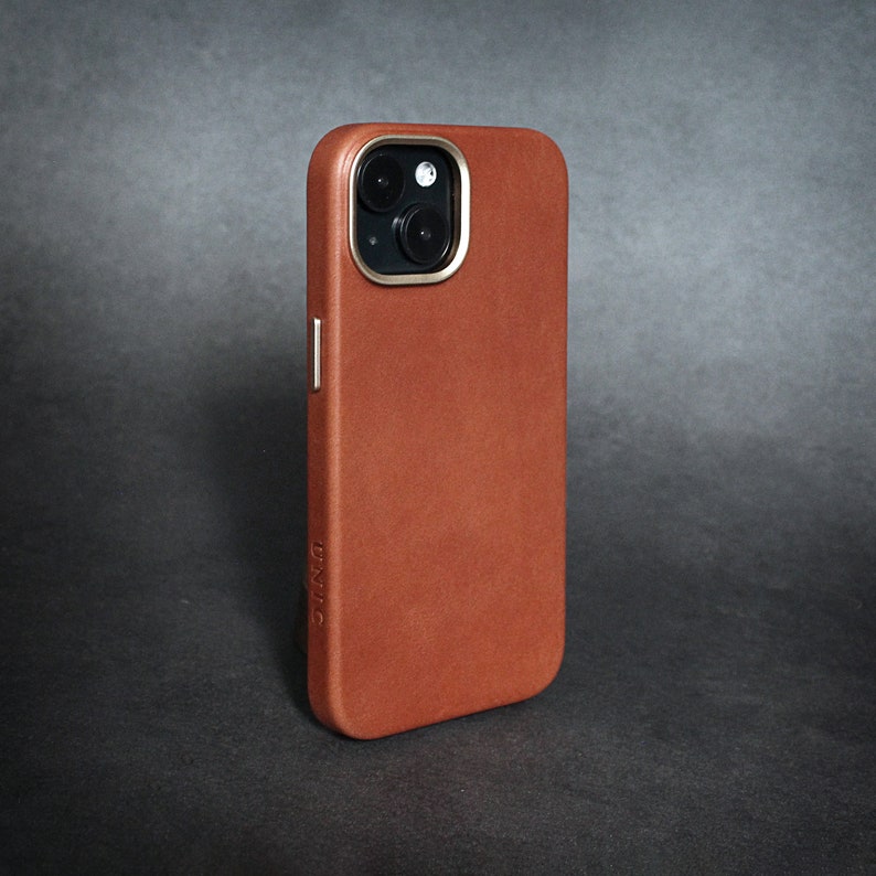 May include: A brown leather phone case with a gold trim around the camera lens. The case has the word "DNC" embossed on the bottom.