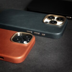 May include: Three phone cases in different colors and materials. The top case is black with a gold camera lens ring. The middle case is dark brown leather with a gold camera lens ring. The bottom case is a light brown leather case with a gold camera lens ring.