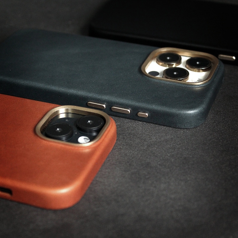 May include: Three phone cases in different colors and materials. The top case is black with a gold camera lens ring. The middle case is dark brown leather with a gold camera lens ring. The bottom case is a light brown leather case with a gold camera lens ring.