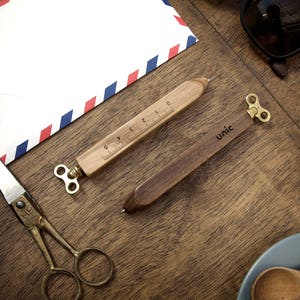 Puede incluir: Dos bolígrafos de madera con un mecanismo de cuerda en forma de llave de latón. Un bolígrafo tiene una regla marcada en centímetros y el otro tiene la palabra "unic" grabada.