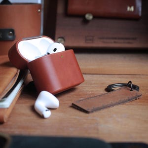 Puede incluir: Una funda de cuero marrón para AirPods con auriculares blancos. Un llavero de cuero a juego está al lado de la funda. Los artículos están sobre una superficie de madera, con otros accesorios de cuero en el fondo.