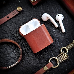 May include: A brown leather case for wireless earbuds. The case has a white interior and a small, black button on the top. The case is sitting on a black leather surface.