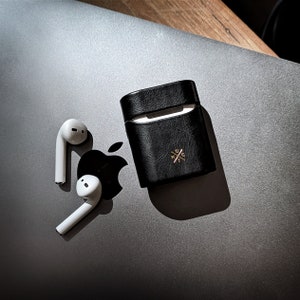 May include: A black leather case for wireless earbuds with a gold cross logo. Two white wireless earbuds are on a silver laptop.
