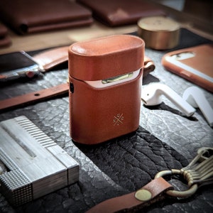 May include: A brown leather case with a white top and a small logo on the front. The case is designed to hold small electronics like earbuds.