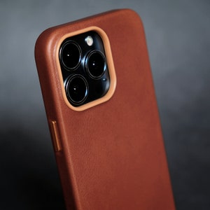 Pu&ograve; includere: Una custodia per telefono in pelle marrone con un ritaglio per l'obiettivo della fotocamera. La custodia ha una superficie testurizzata e una lucentezza sottile.