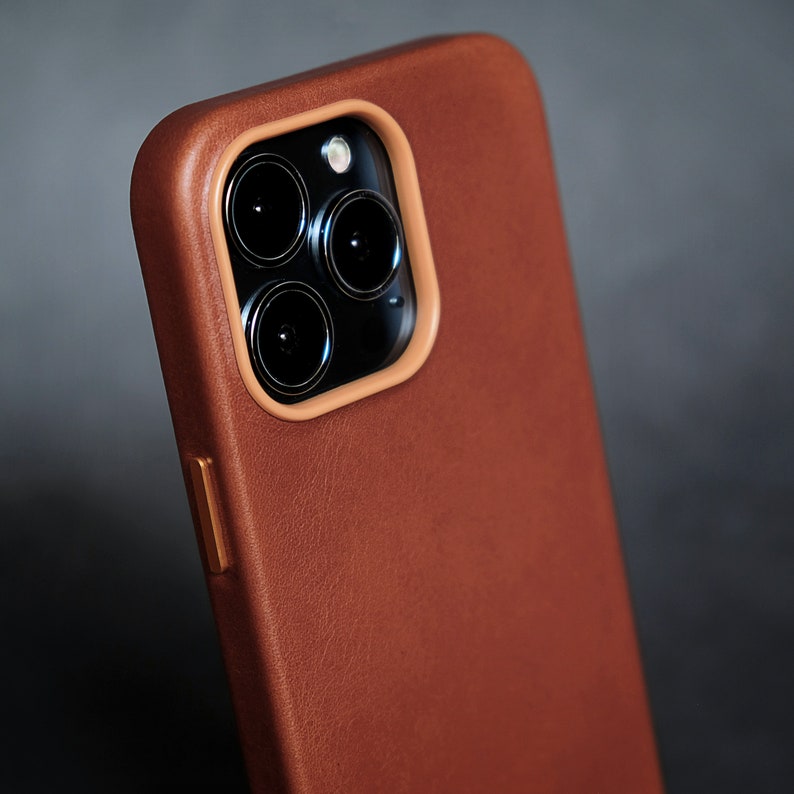Pu&ograve; includere: Una custodia per telefono in pelle marrone con un ritaglio per l'obiettivo della fotocamera. La custodia ha una superficie testurizzata e una lucentezza sottile.
