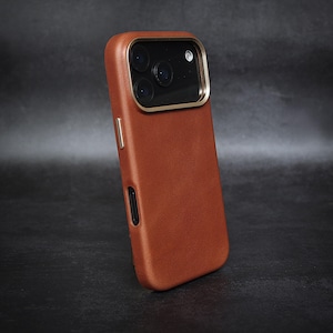 May include: A brown leather phone case with a gold-colored camera frame. The case has a smooth texture and a rectangular shape, with cutouts for the camera lenses and side buttons. The phone case is angled slightly to the left.