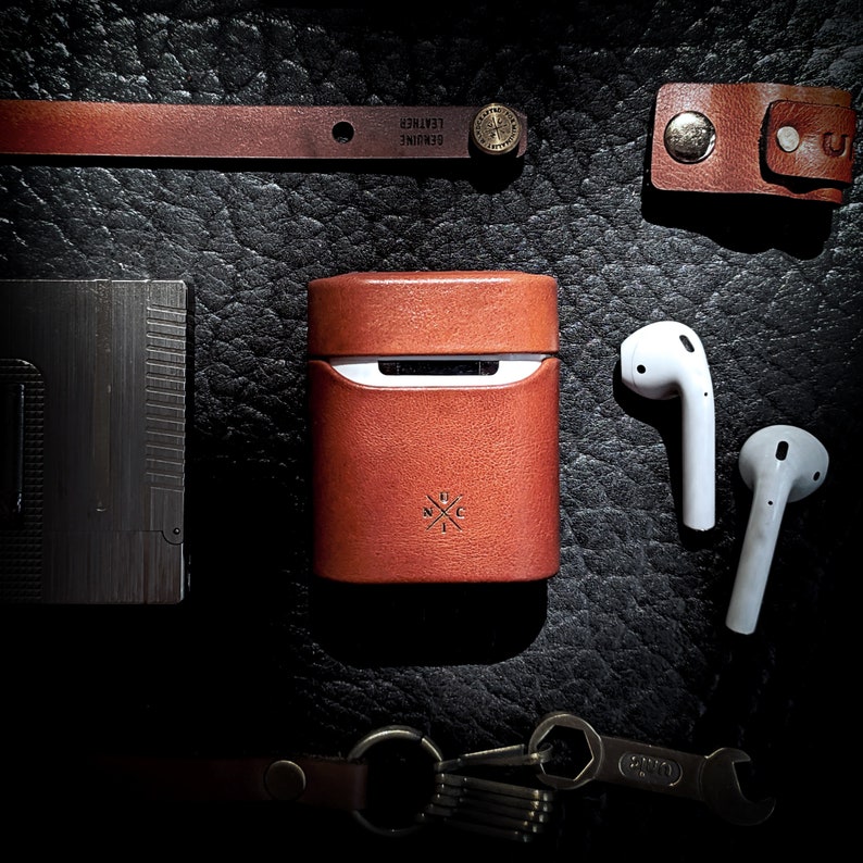 May include: A brown leather case with a metal clasp and the text "Genuine Leather" on the front. The case is open and a white wireless earbud is inside. The case is sitting on a dark brown leather surface.