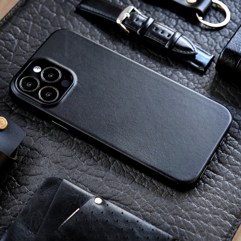Pu&ograve; includere: Una custodia per telefono in pelle nera con un ritaglio per l'obiettivo della fotocamera. La custodia &egrave; appoggiata su una superficie in pelle nera.