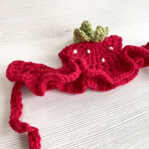 May include: A handmade crochet strawberry headband. The headband is primarily red with white seed details and a green leafy crown. The crochet work is visible, and the headband is designed to be worn around the head.