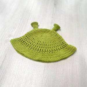 May include: A lime green crochet hat with two small, cylindrical protrusions on top. The hat has a wide brim and a textured, open weave design. The hat is likely handmade and suitable for a child.