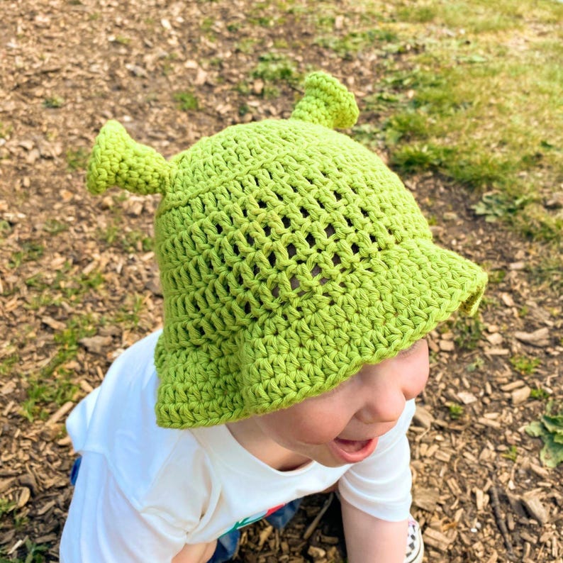 May include: A lime green crochet hat with pointed ears, resembling a character from a popular animated film. The hat has a wide brim and is worn by a child. The hat is made of a loosely woven yarn.