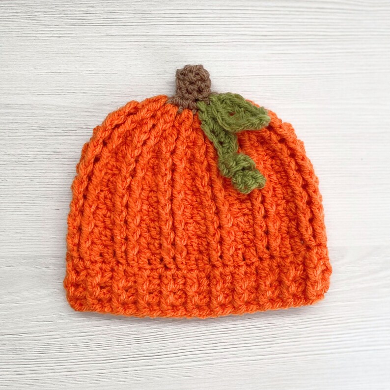 May include: An orange crochet hat shaped like a pumpkin, featuring a ribbed texture. The hat has a brown stem and a green leaf accent, perfect for fall or Halloween. The hat is designed to fit a baby or toddler.