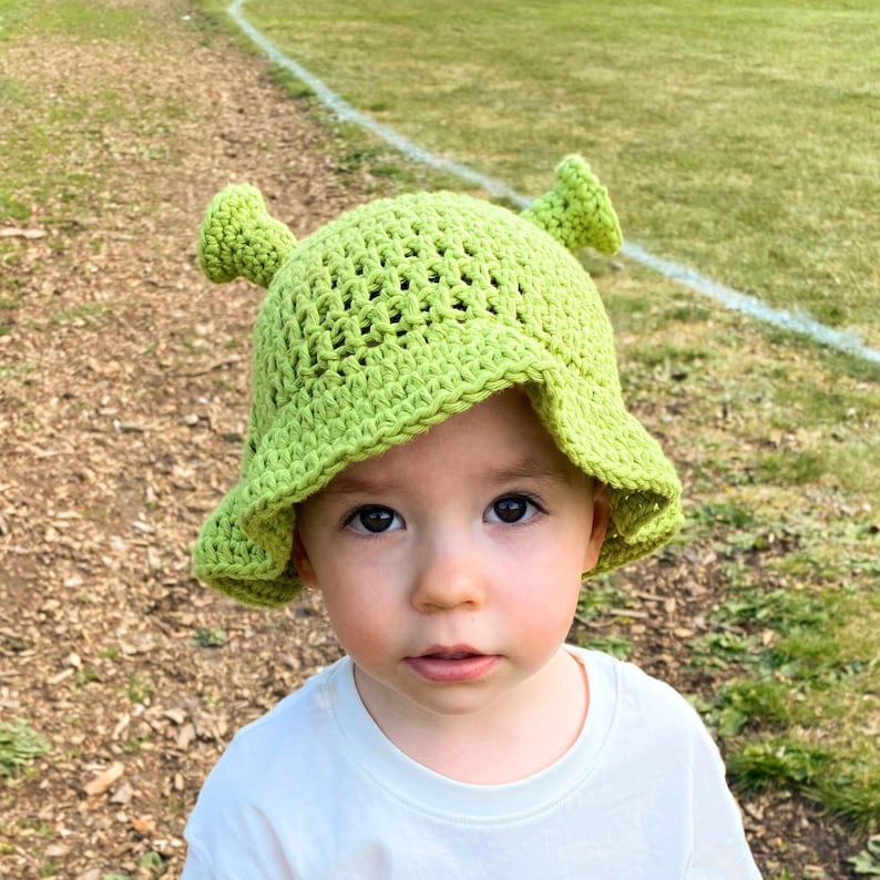 May include: A light green crochet hat with Shrek-like ears. The hat is worn by a young child wearing a white t-shirt. The background is a grassy area with a white line.