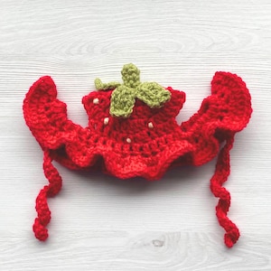 May include: A handmade red crochet strawberry hat with green leaves and white seed details. The hat has a ruffled edge and long, curly ties. This whimsical accessory is perfect for a baby or child.