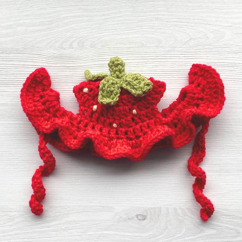 May include: A handmade red crochet strawberry hat with green leaves and white seed details. The hat has a ruffled edge and long, curly ties. This whimsical accessory is perfect for a baby or child.