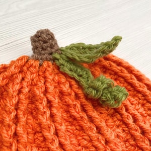 Puede incluir: Primer plano de una calabaza de ganchillo naranja hecha a mano con un tallo marr&oacute;n y hojas verdes. La calabaza est&aacute; hecha con un punto texturizado, ideal para la decoraci&oacute;n de oto&ntilde;o.
