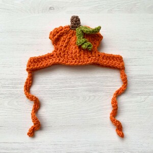 May include: An orange crochet pumpkin hat with a brown stem and green leaves. The hat has long, curly orange ties. This handmade hat is perfect for Halloween or fall-themed events.