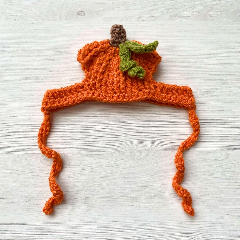 May include: An orange crochet pumpkin hat with a brown stem and green leaves. The hat has long, curly orange ties. This handmade hat is perfect for Halloween or fall-themed events.