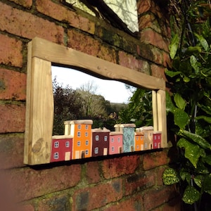 May include: A wooden framed mirror with a wavy glass surface. The mirror is decorated with a row of colorful painted houses.