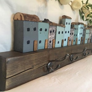 May include: A wooden wall-mounted coat rack with a row of houses painted in shades of blue and gray. The houses are attached to a rustic wooden beam with metal hooks.