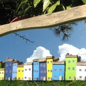May include: A wooden mirror with a curved top frame and a cityscape of colorful houses painted on the bottom edge. The houses are painted in shades of blue, green, yellow, and white.