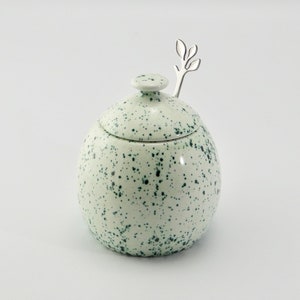May include: White ceramic sugar bowl with a green speckled design and a lid. A small silver spoon with a leaf design is included.