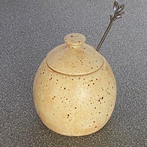 May include: A speckled beige ceramic honey pot with a lid and a small silver spoon with a leaf design.