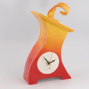 May include: A ceramic clock with a red and yellow gradient glaze. The clock has a white face with black hands and a gold knob. The clock is shaped like a stylized house with a curved top.