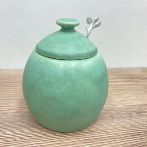 May include: A green ceramic sugar jar with a lid and a small silver spoon with a leaf design.