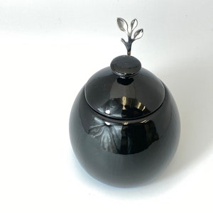May include: A black ceramic jar with a lid and a silver leaf-shaped knob. The jar is round and has a glossy finish.