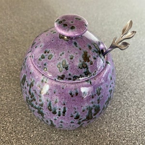 May include: A purple ceramic honey pot with a lid and a small silver spoon with a leaf design. The pot has a speckled glaze.