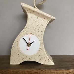 May include: A cream-colored ceramic mantel clock with brown speckles, featuring a curved, asymmetrical silhouette and a decorative spiral handle at the top. The clock face is circular and white with black hour and minute hands and a red second hand.