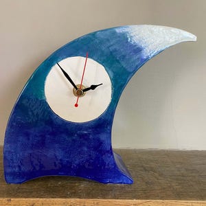 May include: A wave-shaped table clock with a glossy finish in shades of deep blue, teal, and white. The clock face is a circular white disc featuring black hour and minute hands, a thin red second hand, and a central gold-toned metal mechanism.