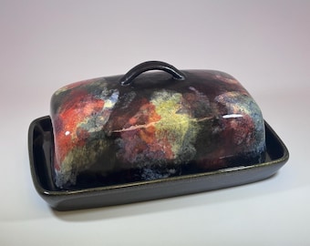 May include: Rectangular ceramic butter dish with a lid. The lid and base are dark brown with a glossy finish. The lid is decorated with a colorful abstract pattern in shades of red, yellow, and blue. The dish has a small, curved handle.