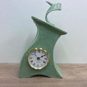 May include: A green ceramic clock with a white face and gold hands. The clock is shaped like a stylized bird with a curved neck and a pointed beak.