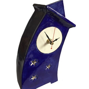 May include: A glossy, deep blue ceramic clock with a curved, asymmetrical silhouette and a peaked top. The clock face is circular and off-white with black hour and minute hands and a thin red second hand. Three star-shaped cutouts decorate the base.