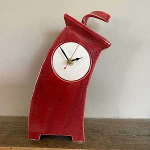 May include: A red ceramic mantel clock with a curved, leaning silhouette and a speckled glaze finish. The clock face is circular, white, and features black hour and minute hands with a thin red second hand, all centered on the front surface.