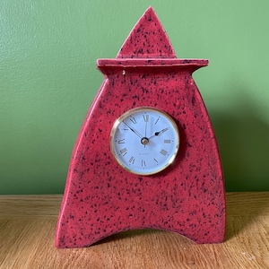 May include: A red ceramic clock with black speckles in the shape of a house. The clock face is white with black Roman numerals and gold hands. The clock says "Quartz" on the face.