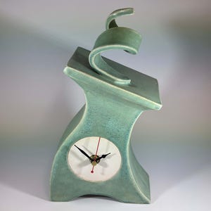 May include: A sage green ceramic clock with a curved, asymmetrical body and a smooth, matte finish. The clock face is circular, white, and features black hour and minute hands with a thin red second hand, centered on the lower front section.
