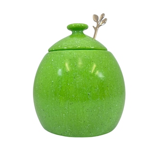 Könnte beinhalten: Ein grüner, gesprenkelter Keramik-Honigtopf mit Deckel und einem kleinen silbernen Löffel mit Blattdesign.