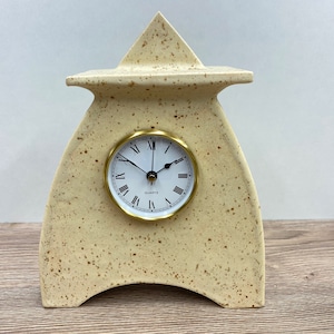 Puede incluir: Un reloj de cerámica beige con una esfera dorada y números romanos. El reloj tiene forma de casa con un techo puntiagudo.
