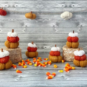 Può includere: Cinque zucche all'uncinetto in arancione, bianco e marrone, impilate in gruppi di tre e due, con una spolverata di mais dolce intorno.