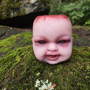 May include: A hollow doll head planter with pale skin, visible blue veins, and dark red shading around the eyes and top rim. The doll features light brown eyebrows, grey eyes, and a slightly open mouth showing small teeth, with a flat open top.
